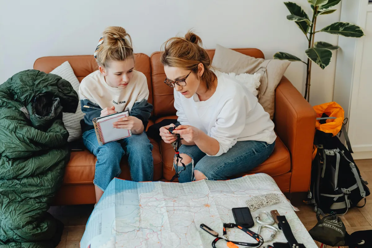Two individuals sit on a couch, examining a map and outdoor gear, preparing for an adventure. A sleeping bag and backpack are nearby.