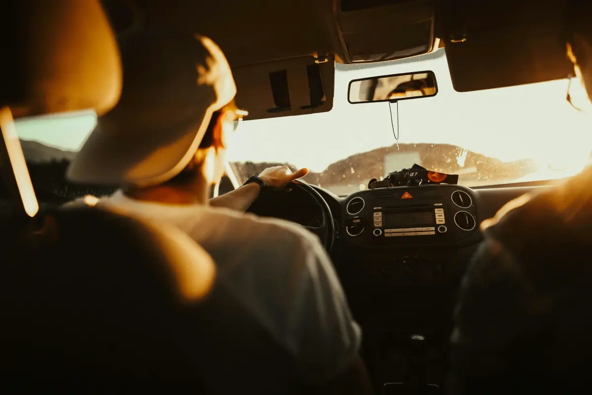 A person drives a car during sunset, with sun rays illuminating the scene and a passenger visible in the foreground.