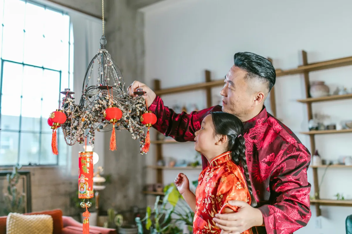 A person in a traditional red outfit adjusts a decorative chandelier with red lanterns in a cozy, modern home setting.