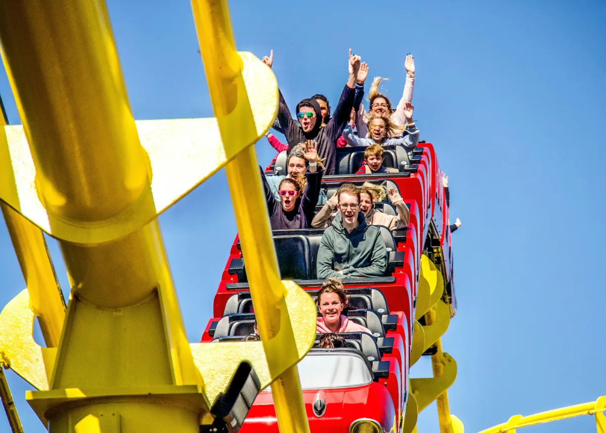 A red roller coaster climbs against a clear blue sky, with excited riders raising their hands in joy.