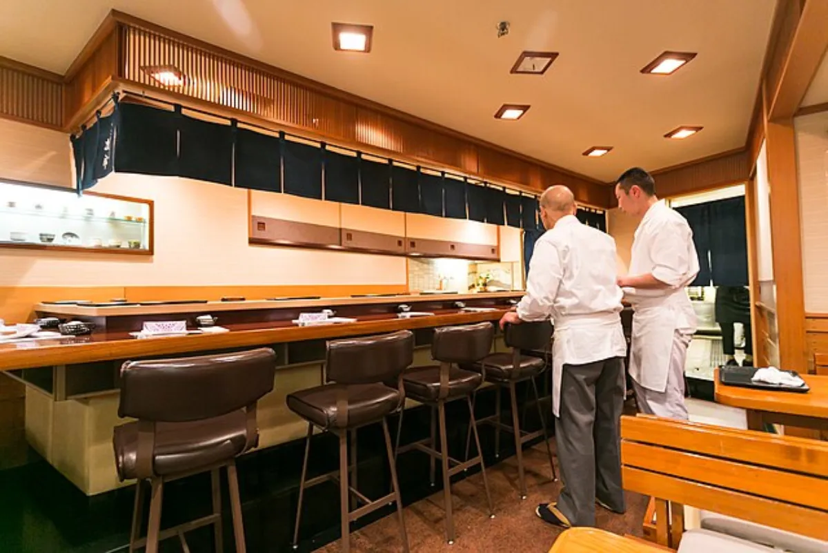 Two chefs in white uniforms stand in a cosy restaurant kitchen, surrounded by bar stools and a wooden counter.
