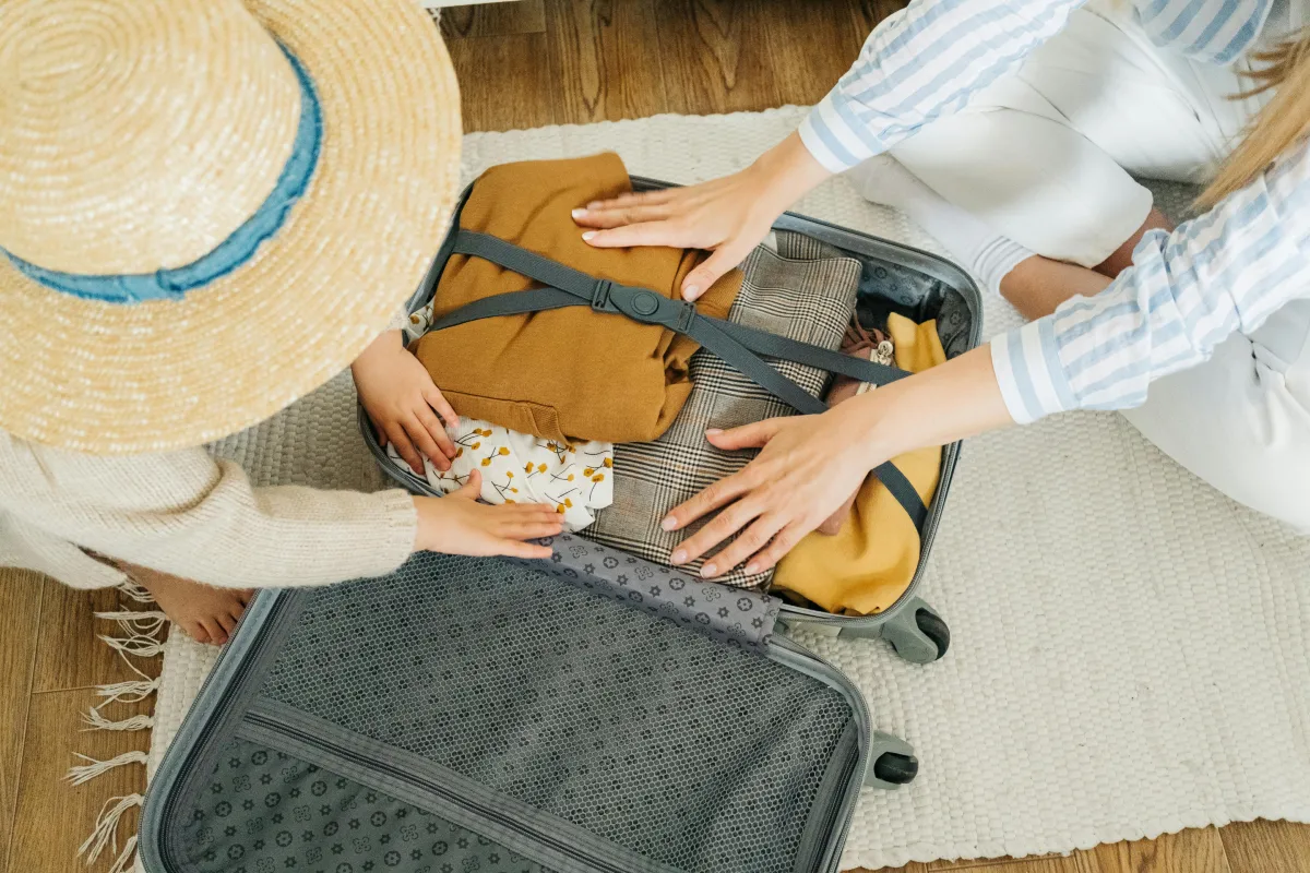 A child in a straw hat helps an adult pack a suitcase filled with various clothes on a wooden floor.