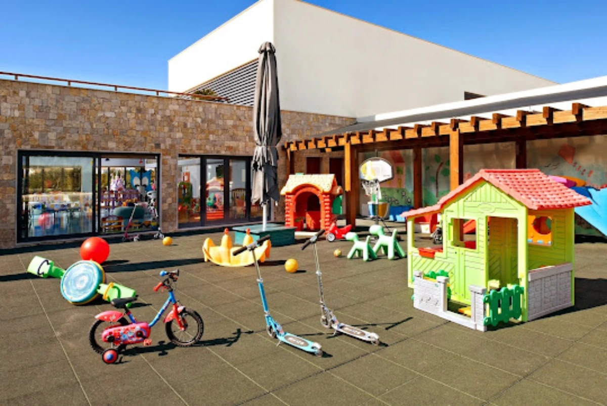 Colorful outdoor play area featuring bikes, scooters, playhouses, and balls on a sunny day, with a stone wall and shaded seating.