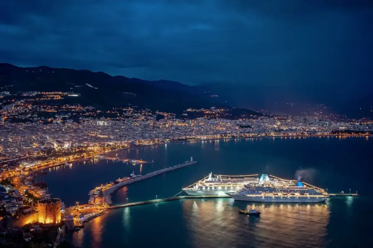 Aerial view of a coastal city at dusk, featuring illuminated buildings and a busy harbour with a cruise ship.