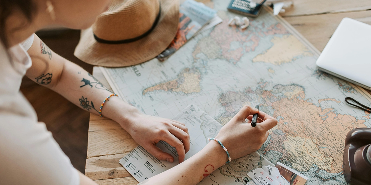 A person with tattoos writes on a world map, surrounded by travel documents and a hat, planning their next adventure.