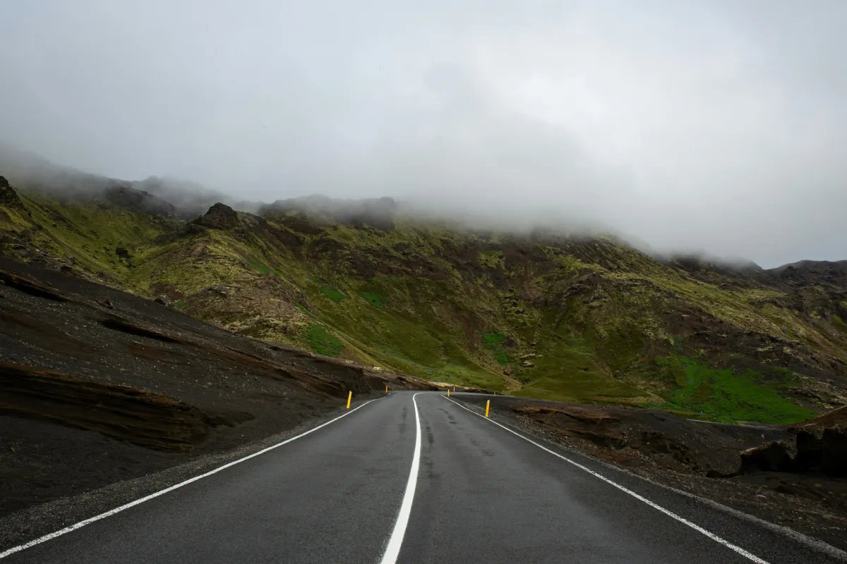 A winding road stretches through lush green hills, partially shrouded in fog, under an overcast sky.