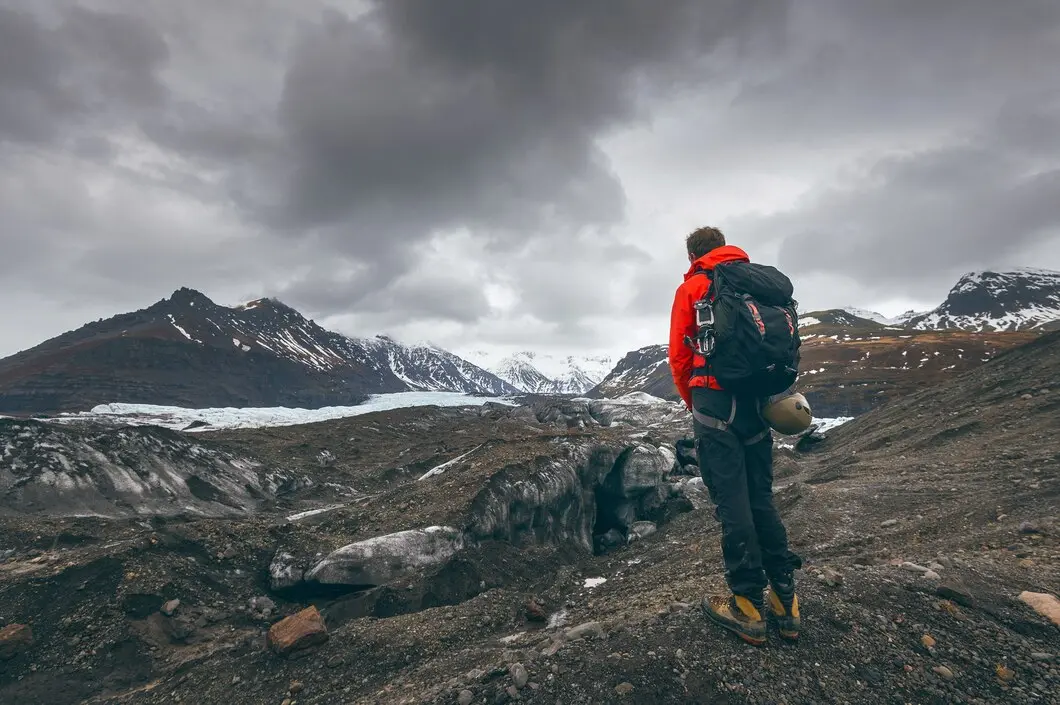 A hiker in a bright red jacket stands on a rocky glacier landscape, gazing at distant mountains under a cloudy sky.