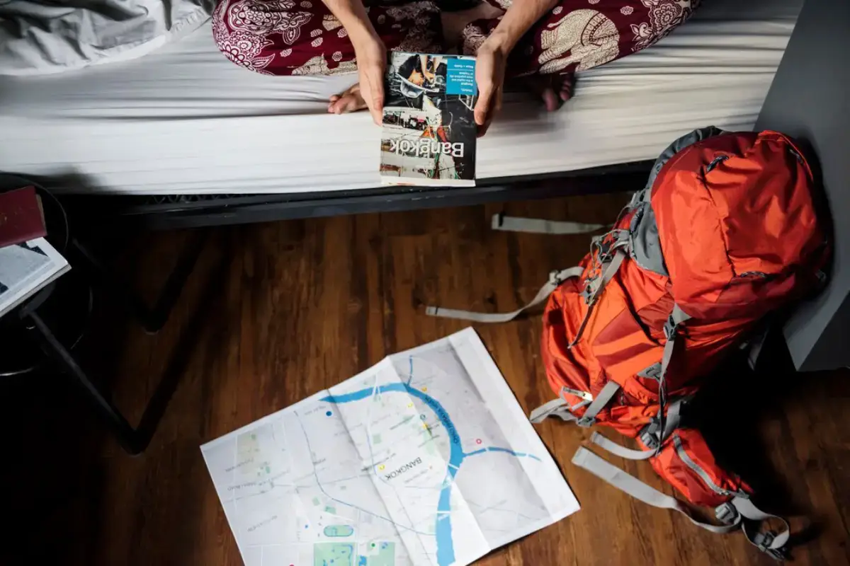 A person sitting on a bed, holding a travel guide, with a colourful backpack nearby and scattered books on the bed.
