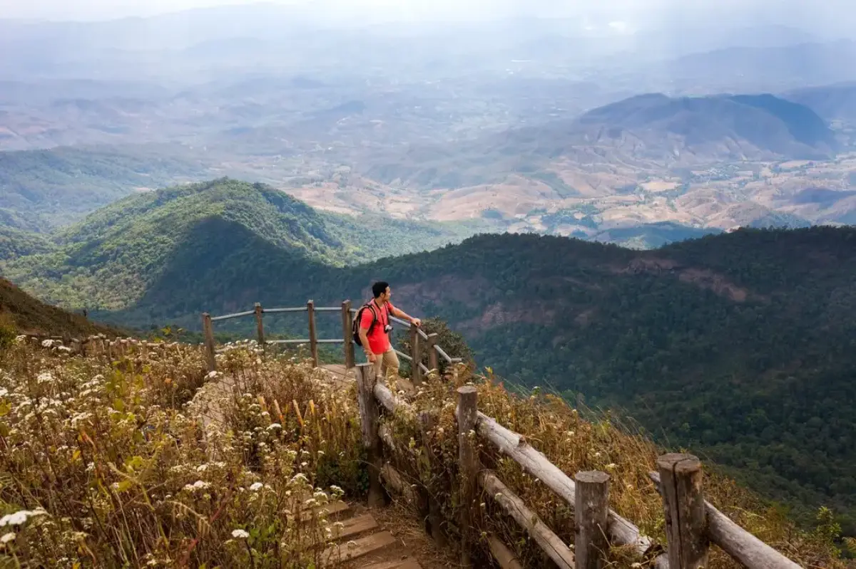 A hiker in a red shirt stands on a wooden path overlooking lush green mountains and valleys under a cloudy sky.