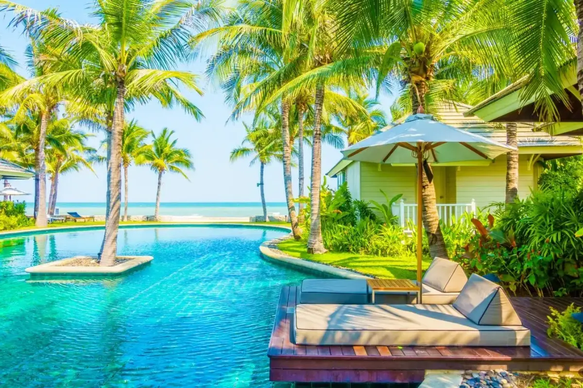 A serene pool, lush palm trees, and a cosy lounge area overlooking the ocean on a sunny day.