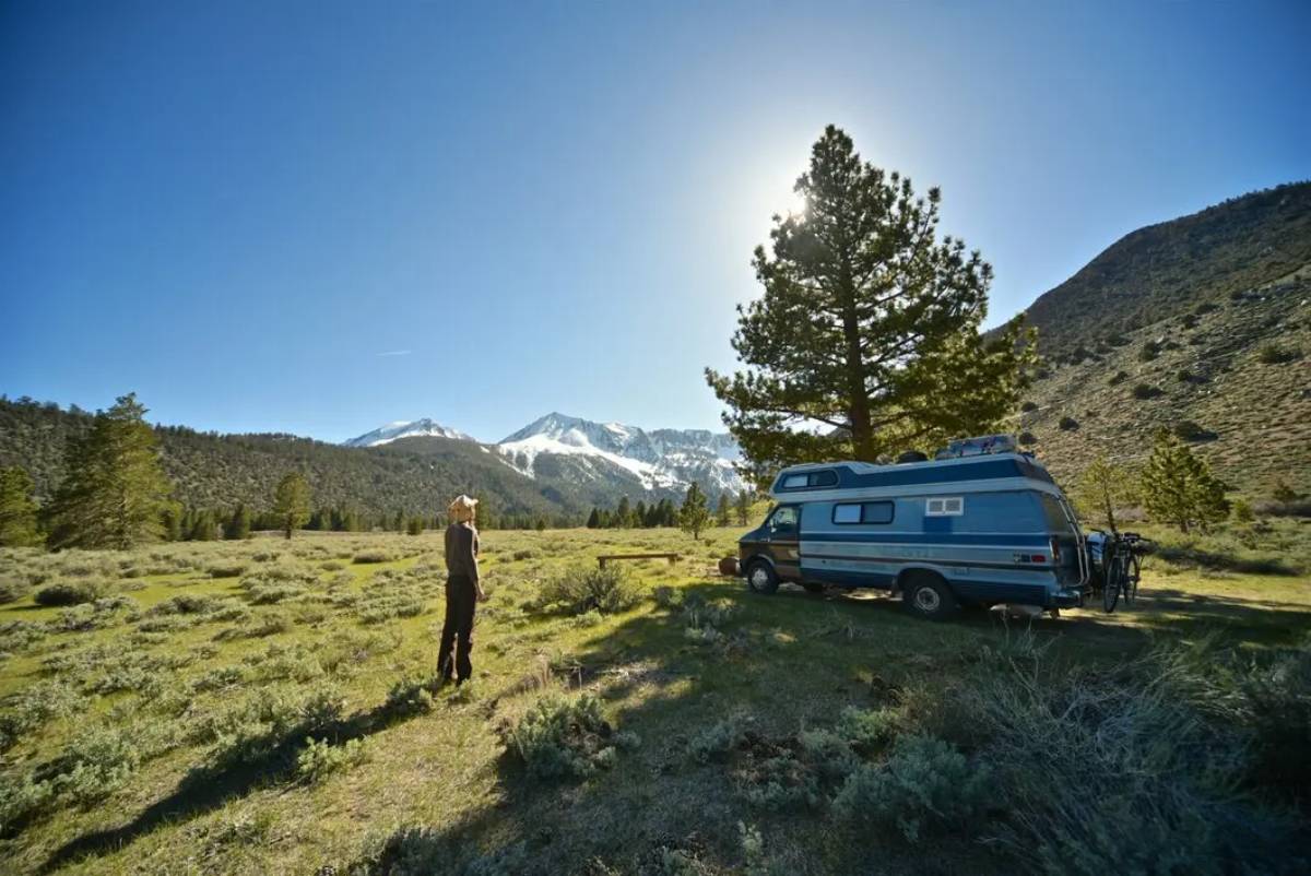 A person stands near a vintage camper van in a serene mountainous landscape with snow-capped peaks and clear blue skies.