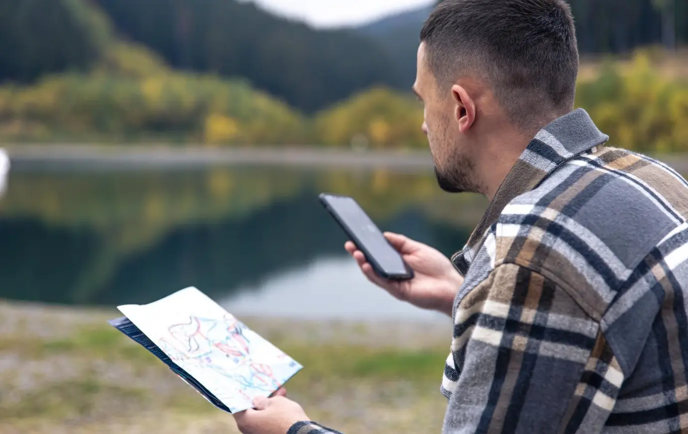 A person holds a map in one hand and a smartphone in the other, seated by a serene lake surrounded by autumn foliage.