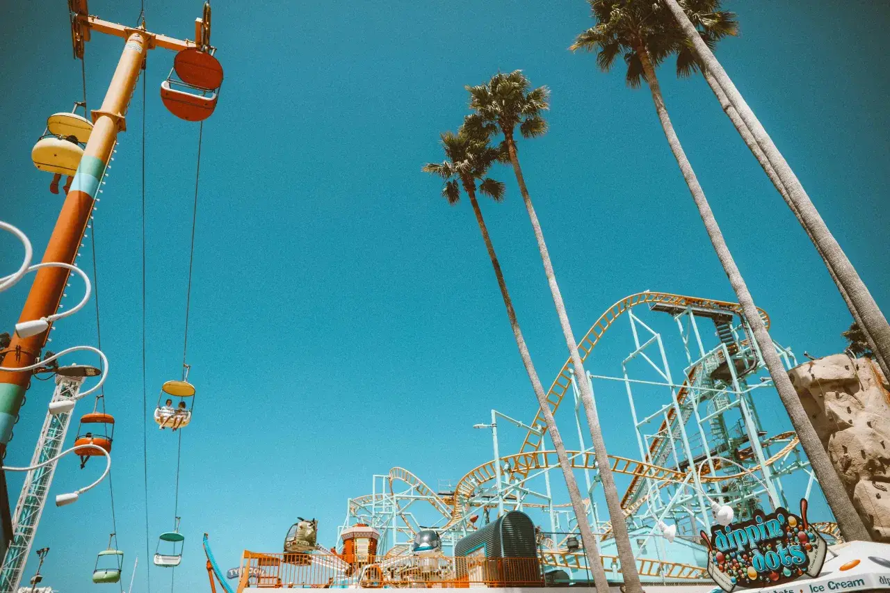 Bright blue sky above a lively amusement park with palm trees, a roller coaster, and colorful gondolas on a cable car ride.