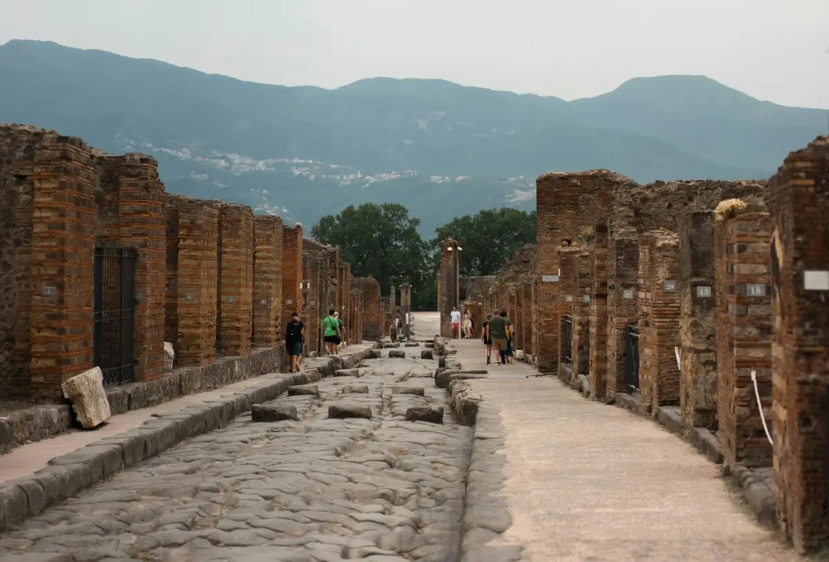 Ruins of ancient Pompeii with tourists walking along cobblestone streets, framed by weathered brick walls and distant mountains.