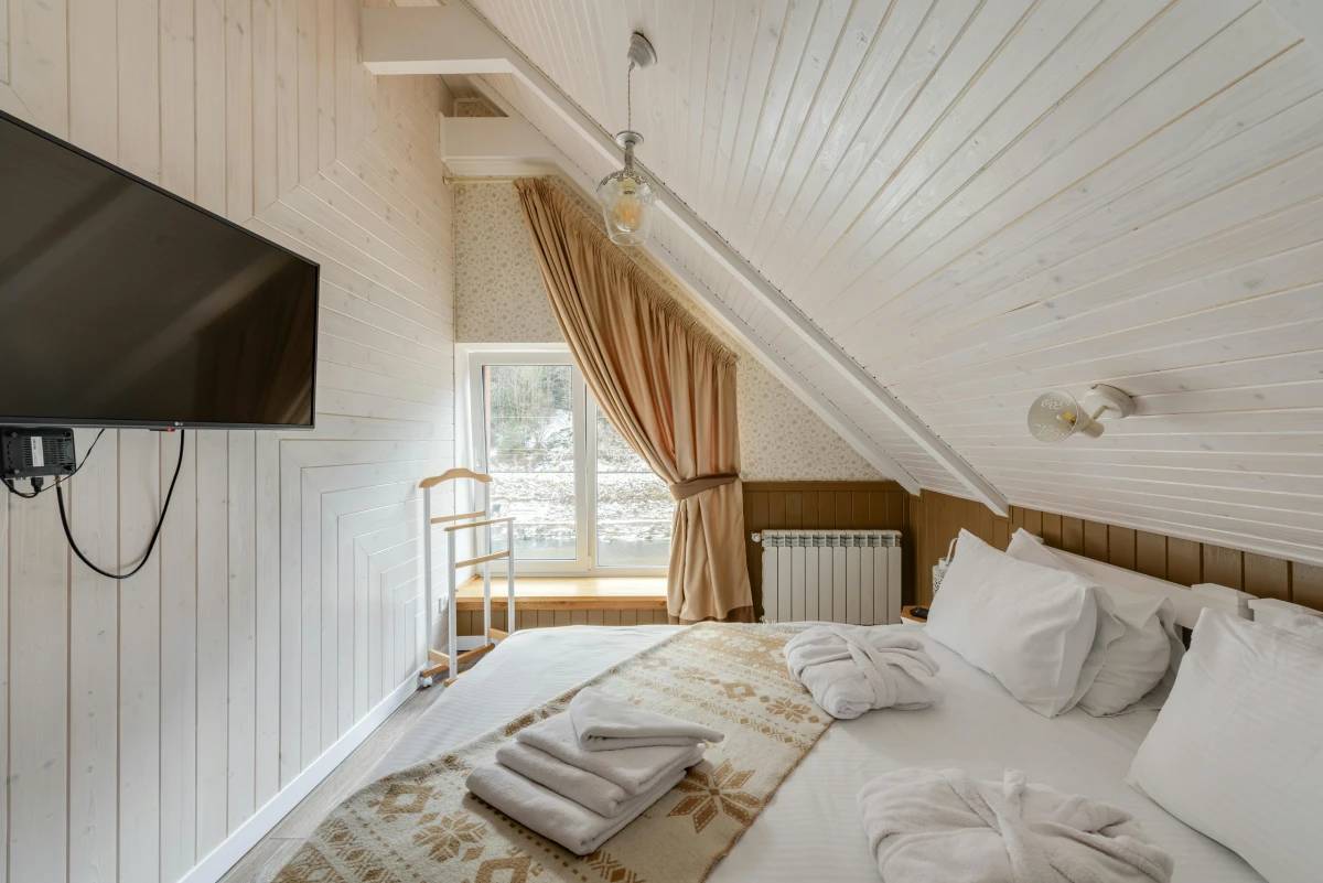 Cozy attic bedroom with white wooden walls, a large window with curtains, a flat-screen TV, and a neatly made bed with towels.
