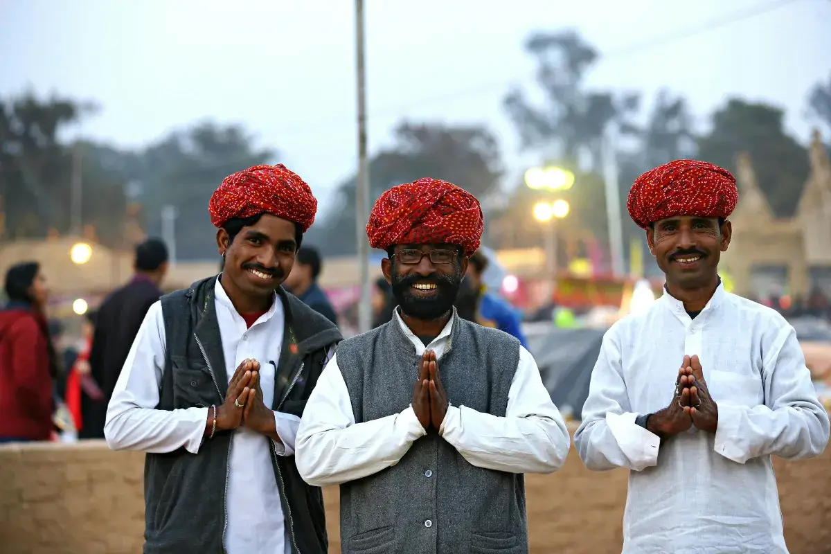 Three men in traditional Rajasthani attire with red turbans standing in prayer pose against a softly lit outdoor background.