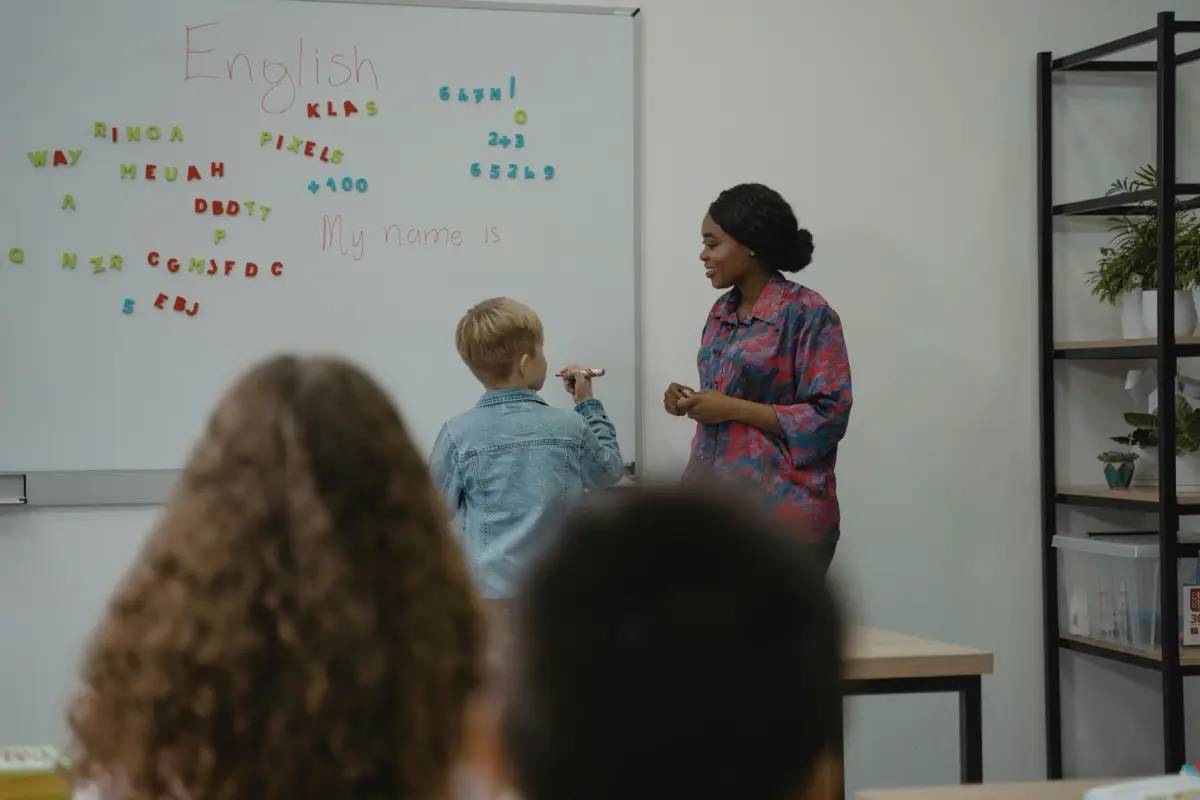 A teacher stands beside a whiteboard filled with colorful letters and words, while a student writes using a marker.