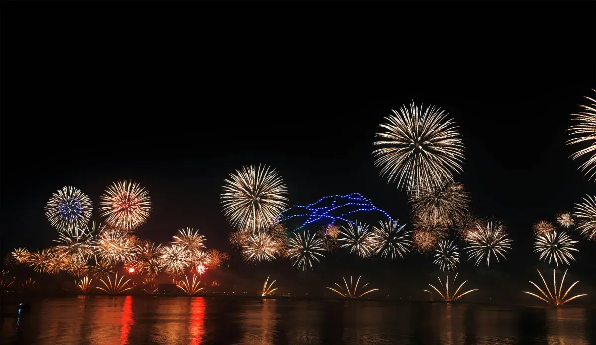Colorful fireworks illuminate the night sky above a body of water, with a brightly lit structure in the background.