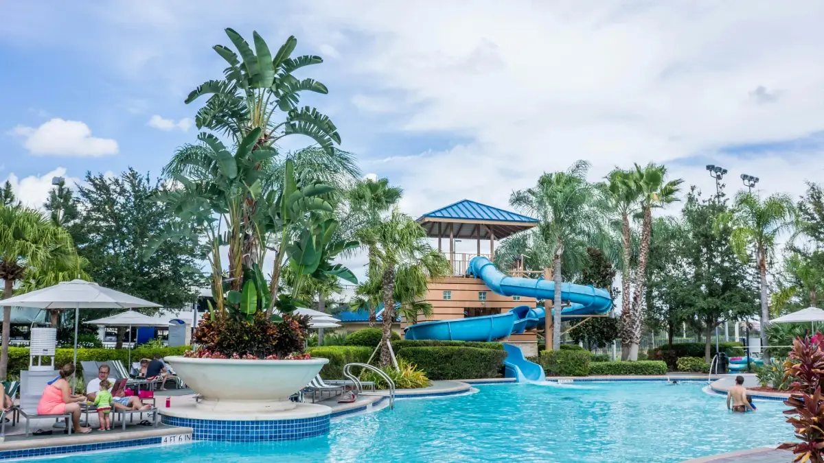 A bright outdoor pool area featuring a blue waterslide, tropical plants, and a relaxing atmosphere under a cloudy sky.