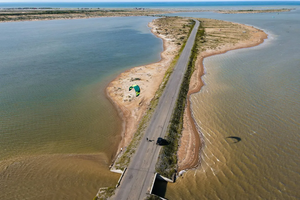 Aerial view of a narrow road along a coastline, with a kite surfer nearby, surrounded by tranquil waters and sandy shores.