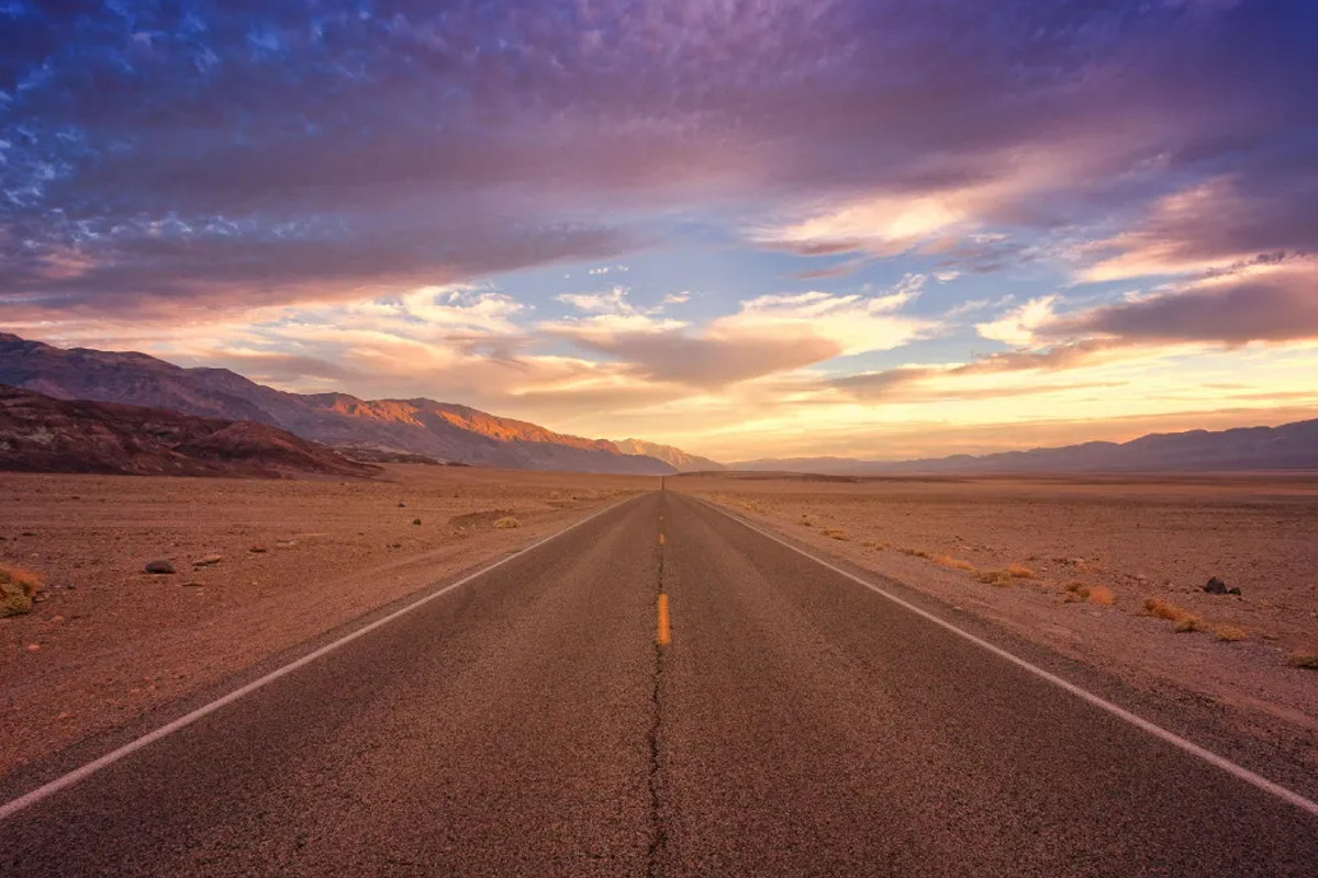 A long, straight road stretches through a desert landscape, framed by mountains under a colorful sunset sky.